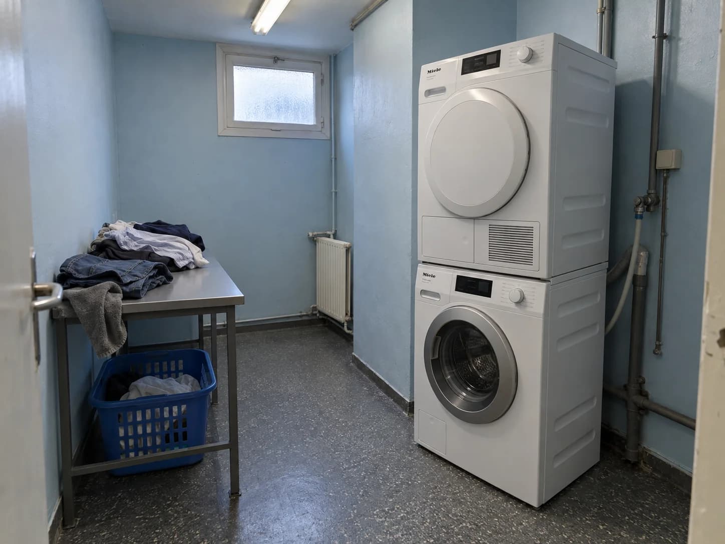 Lave-linge et sèche-linge en colonne dans un petit local d’internat aux murs bleus, avec une table pour plier le linge